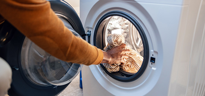 A person is loading laundry into a front‑loading washing machine. Their hand is pushing a pile of striped fabric into the drum. The washer door is open, showing the metallic interior and rubber gasket around the rim. The scene suggests starting a wash cycle in a home laundry area.