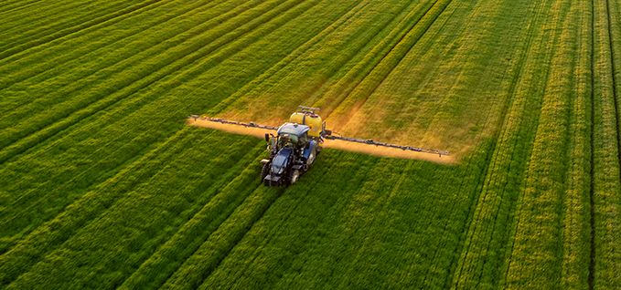 Aerial view of a tractor spraying crops across a large green field at sunset.