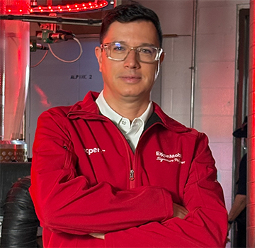Person wearing a red ExxonMobil jacket with arms crossed in an industrial setting with metal pipes and red lighting.