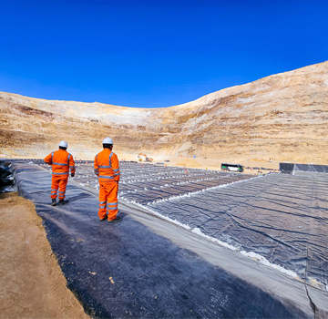 Two workers in orange safety gear at a lined open-pit site under a blue sky.